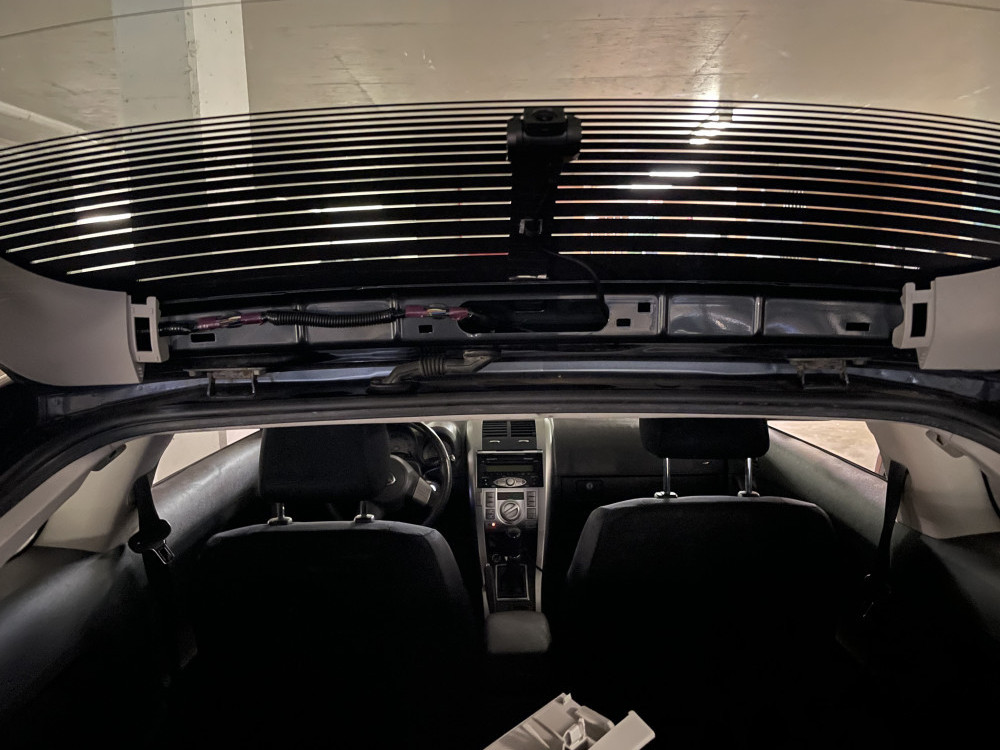 Rear dash cam mounted on hatchback door with piece of metal wedged between rear glass and metal door frame.