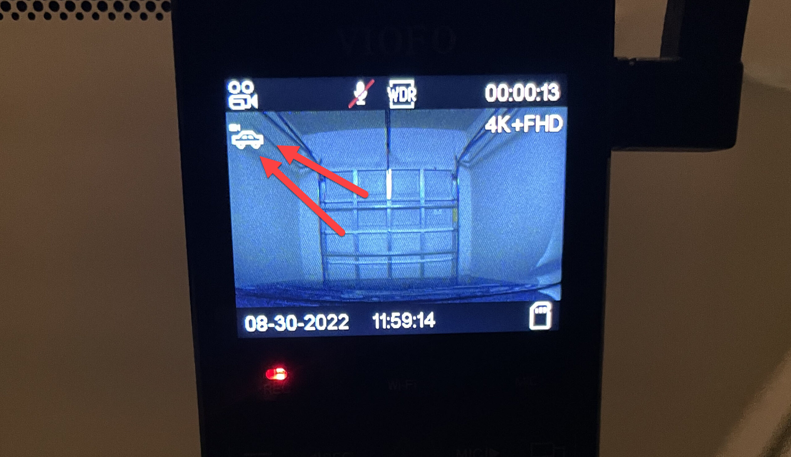 A picture of the screen of a dash cam with two red arrows pointed towards the the regular driving mode icon. There is a closed garage door on the dash cam screen.